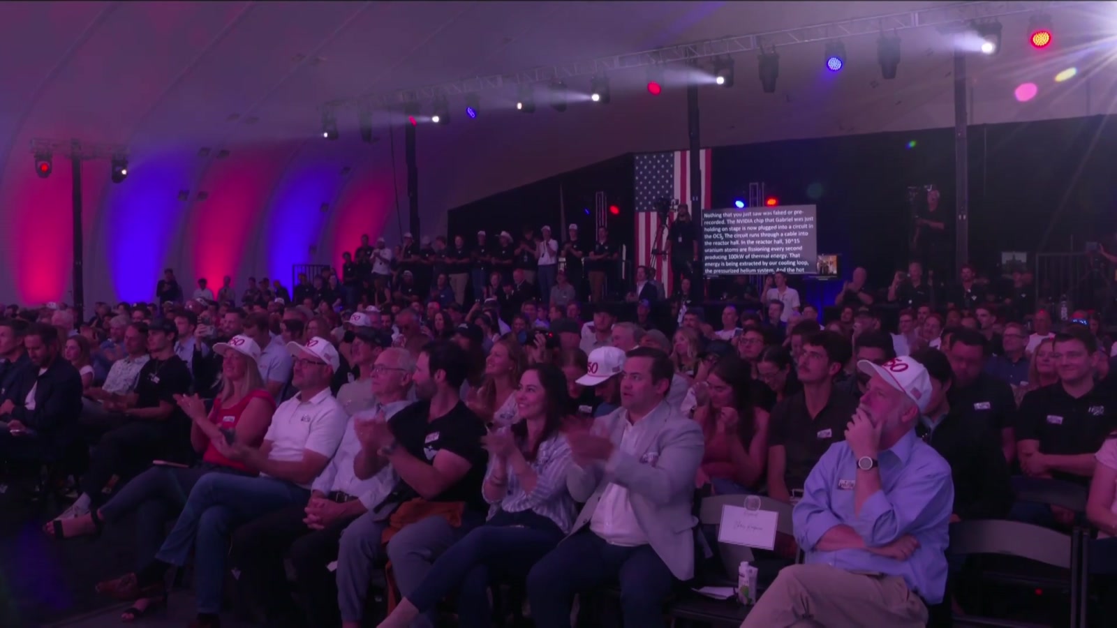 Ein voll besetzter Veranstaltungsraum mit blau-rotem Bühnenlicht, das Publikum applaudiert in Richtung Bühne.